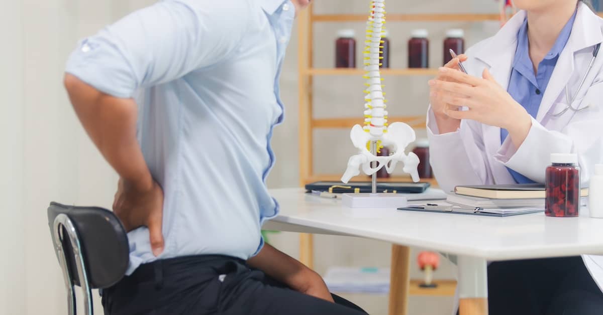 Doctor pointing out spinal injuries on a model of a spine to a patient with back pain | Burg Simpson